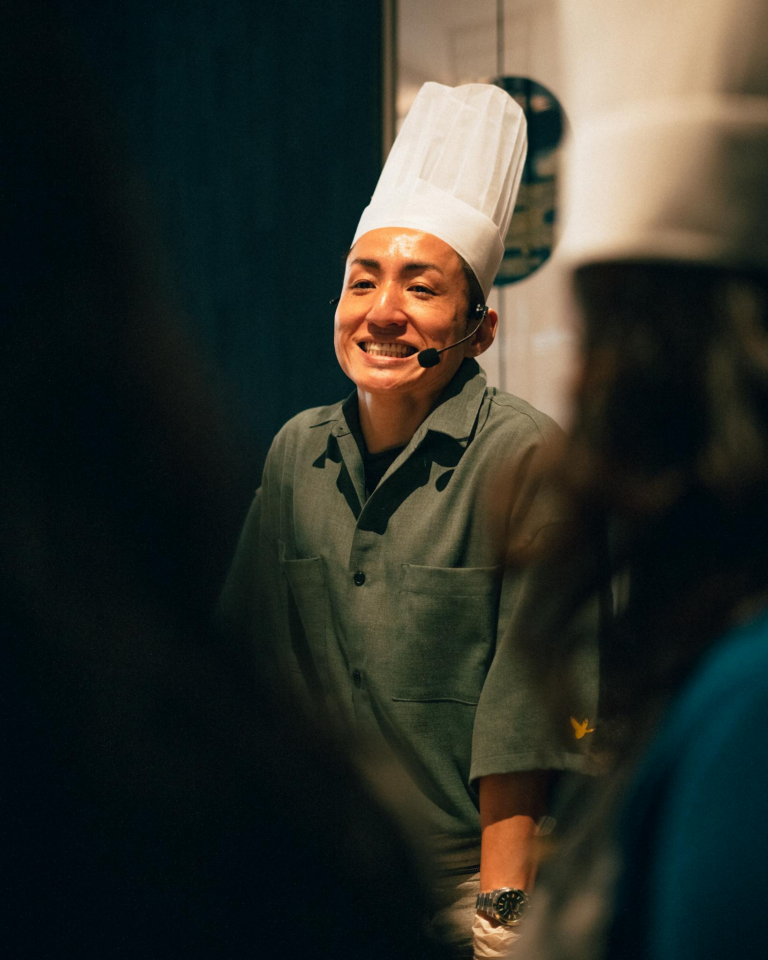 Happy chef wearing a hat and microphone, delivering a cooking presentation indoors.
